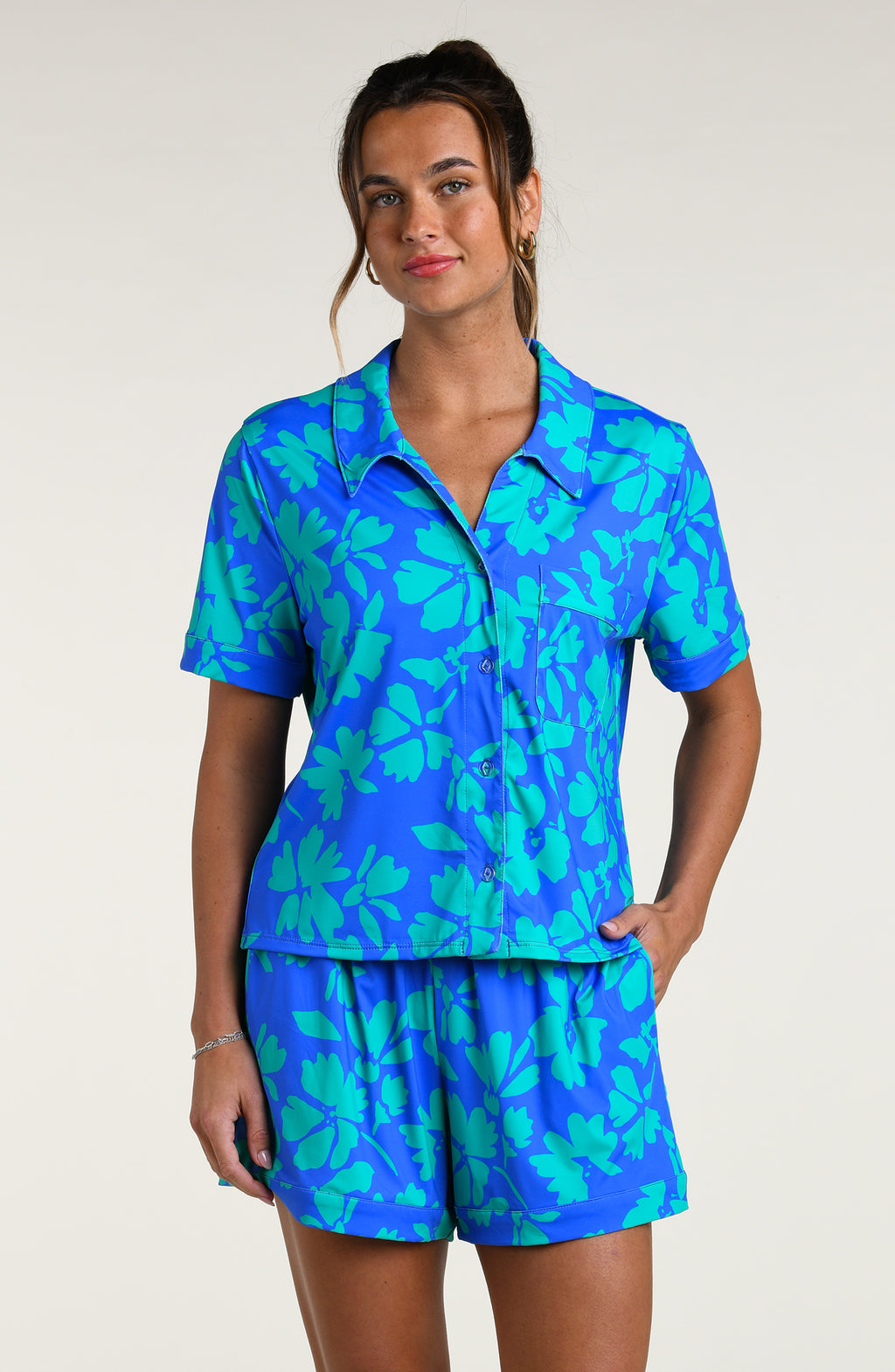 Woman wearing a blue and green floral patterned short sleeve shirt and shorts pajama set against a white background
