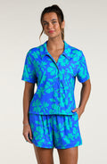 Woman wearing a blue and green floral patterned short sleeve shirt and shorts pajama set against a white background