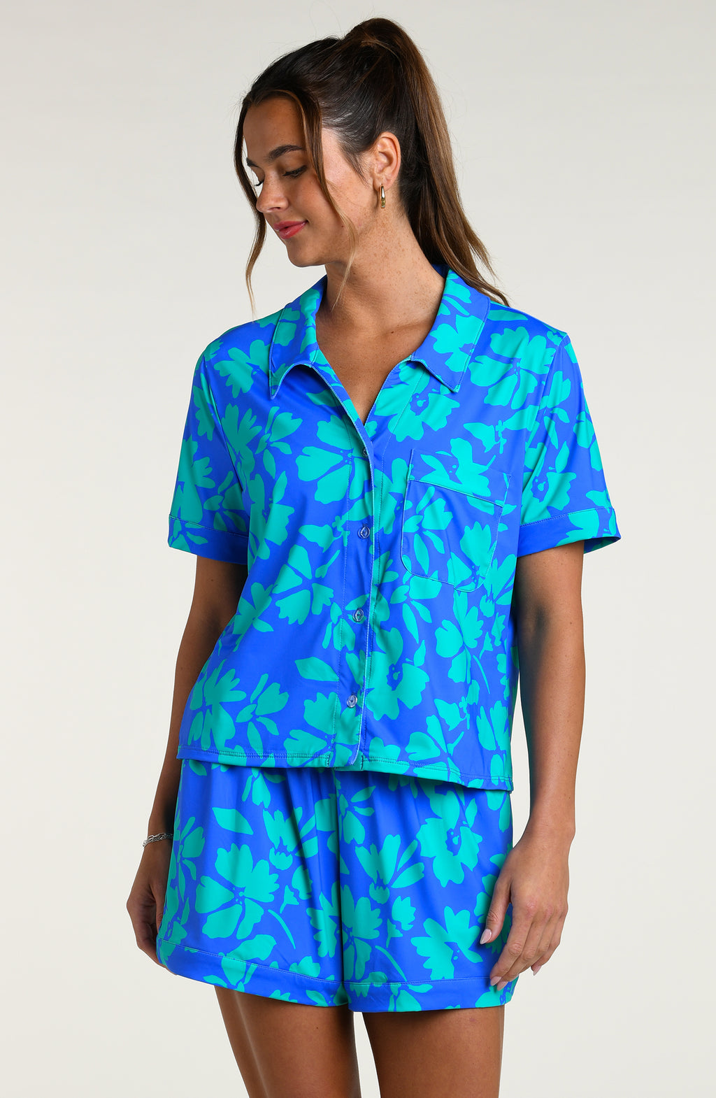 Woman wearing a blue and green floral patterned short sleeve shirt and shorts pajama set against a white background