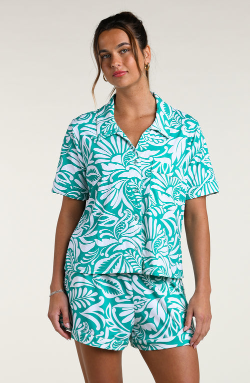 Woman wearing a teal and white patterned short sleeve shirt and shorts pajama set against a plain background