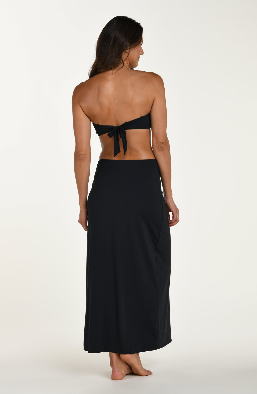 Full body back image of model wearing a black swim skirt with ruched side detail and a high slit on one leg. This is paired with a black bikini top.