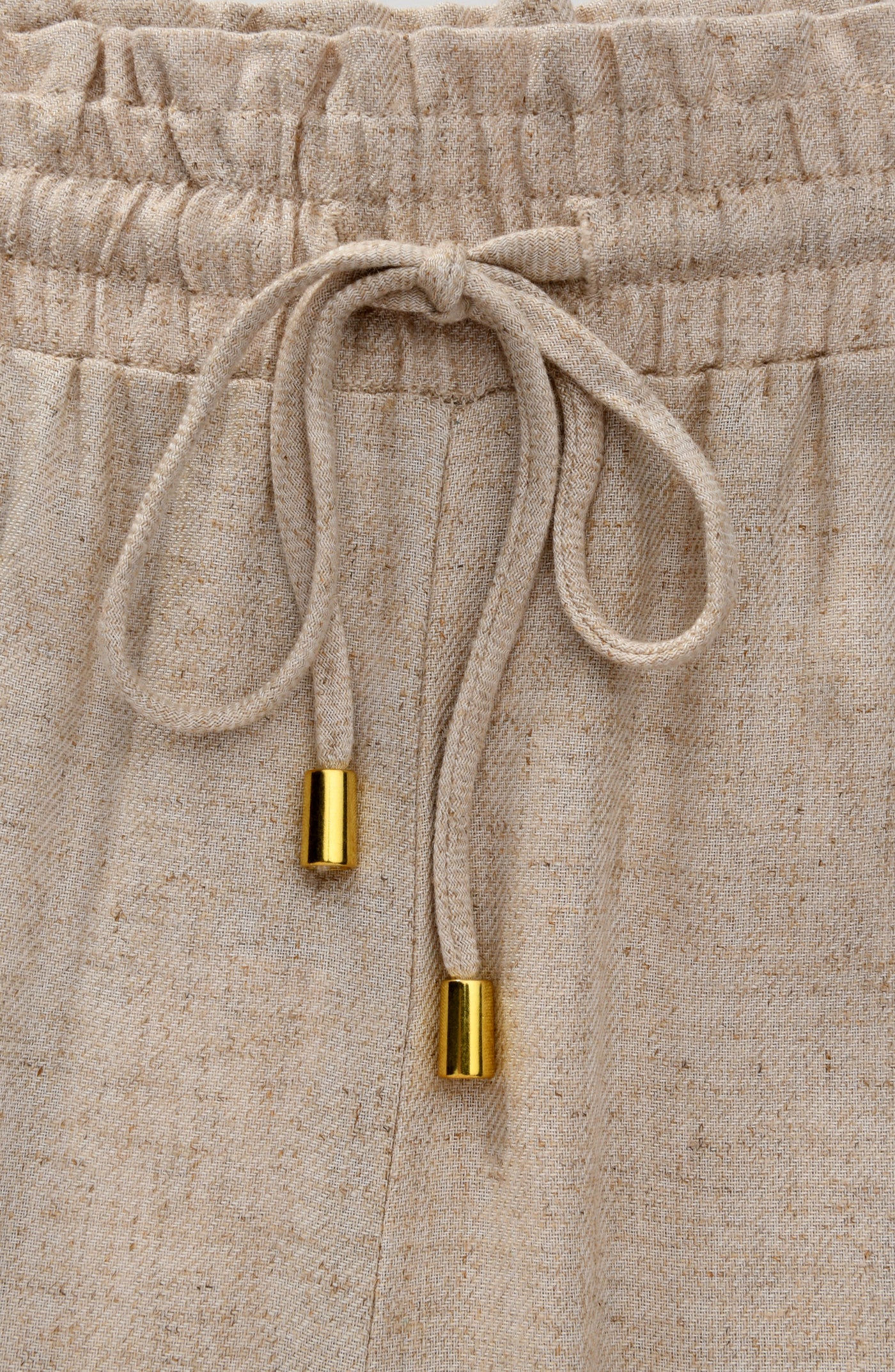 
                  
                    Beige pants with a drawstring and gold accents on a matching background
                  
                