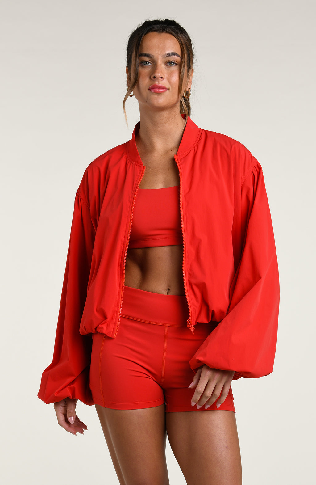 Woman wearing a red athletic jacket over a red sports bra and matching bike shorts against a white background