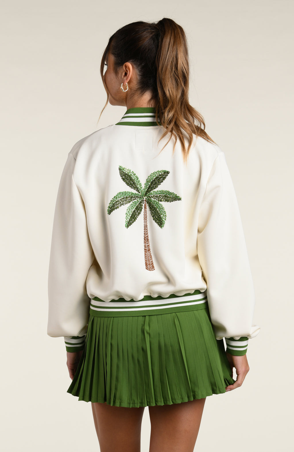 Person wearing a white varsity jacket with a palm tree design and green pleated skort on a plain background