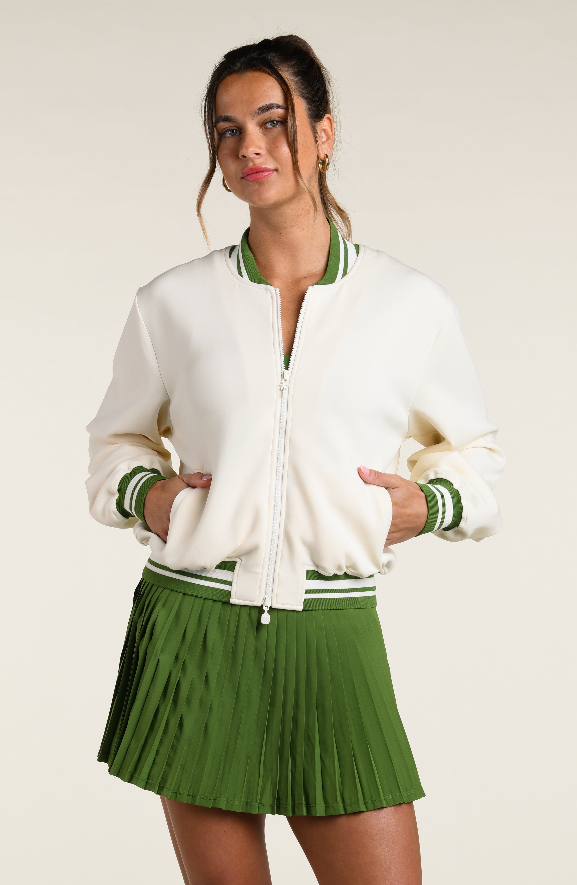 Woman wearing a white varsity jacket with green trim and a green pleated skort on a plain background