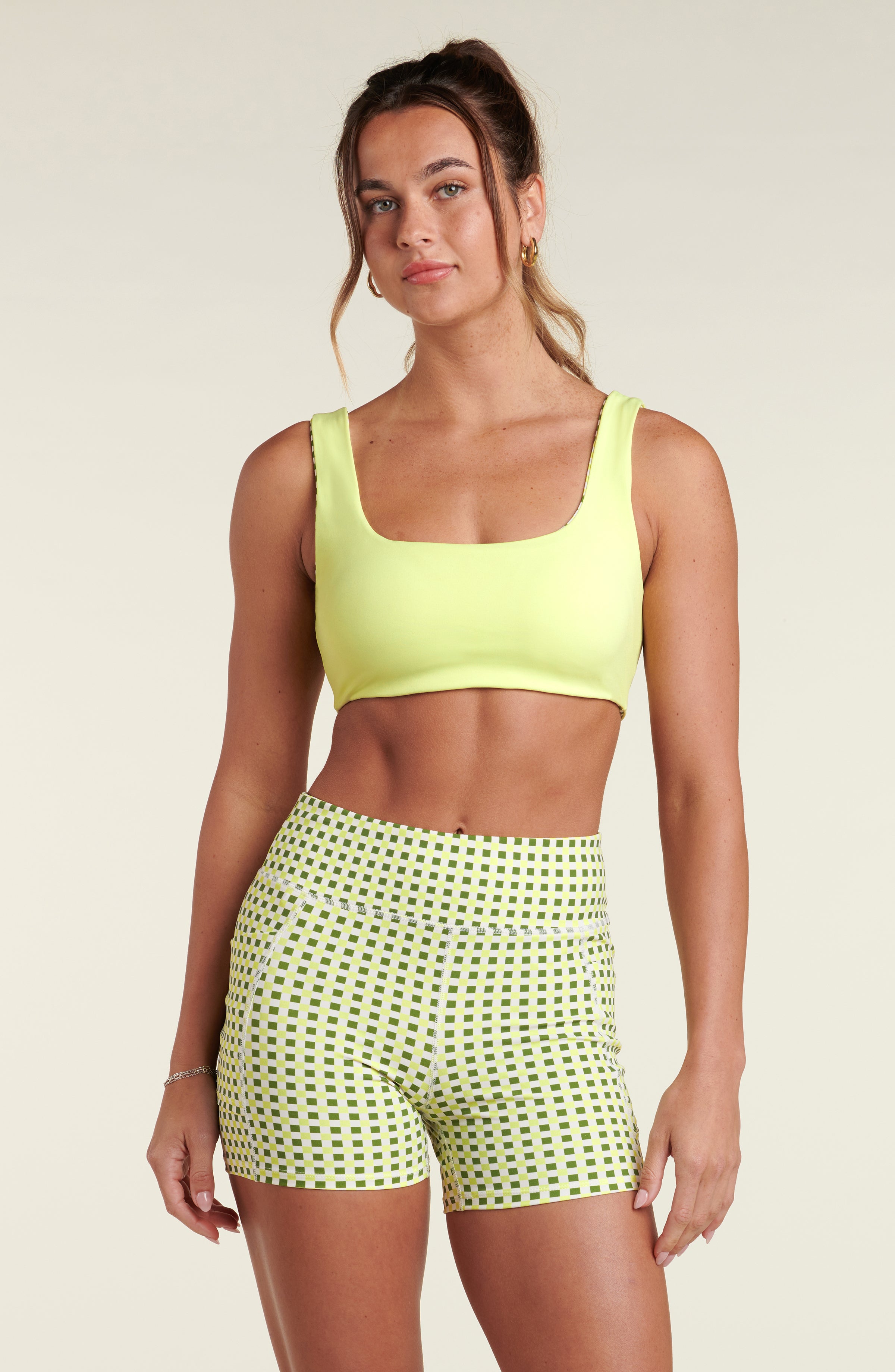 Woman wearing a lime green sports bra and green checkered biker shorts against a beige background