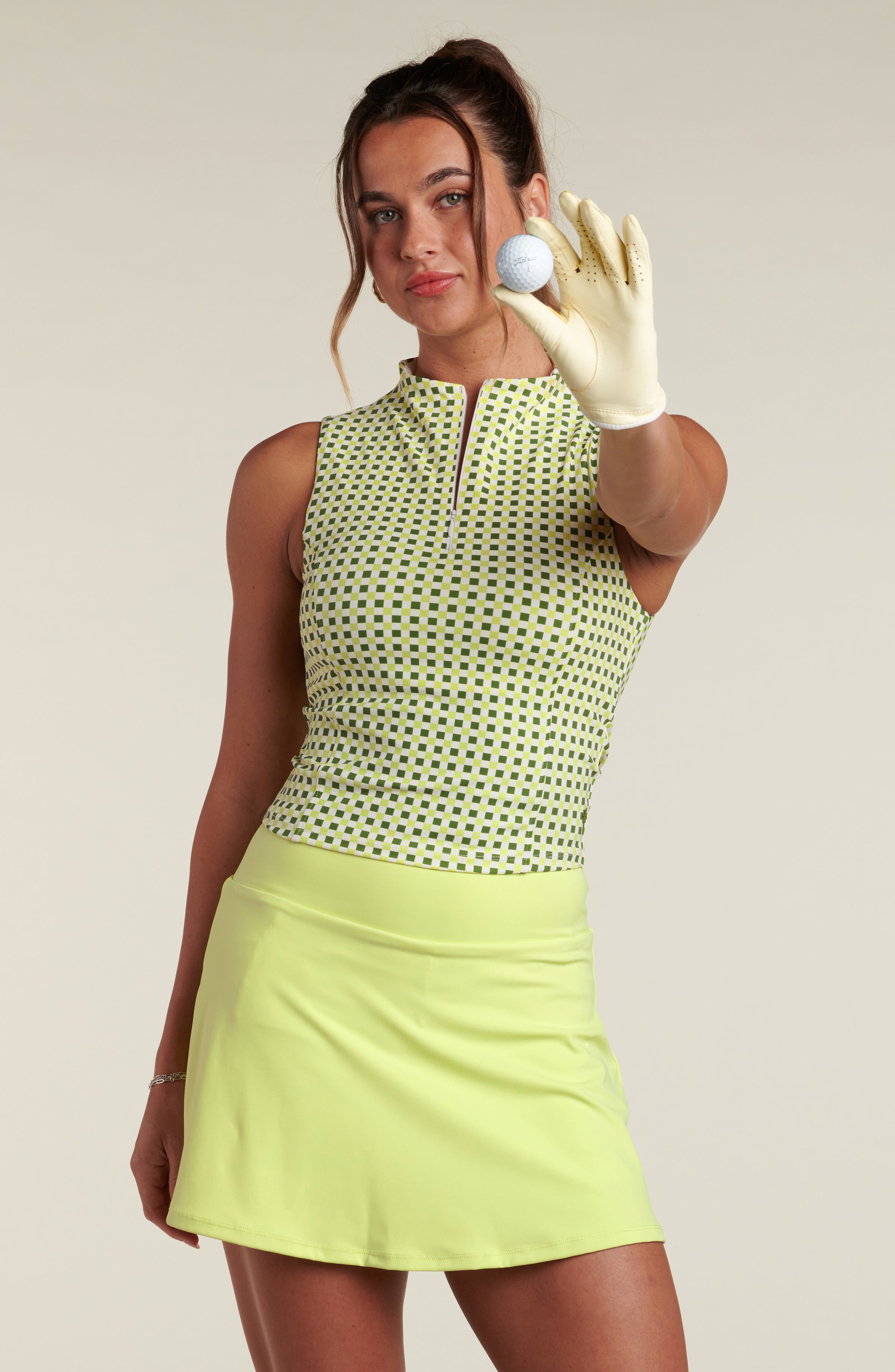 Woman wearing a green patterned sleeveless top and lime green skirt against a beige background, holding a golf ball. 