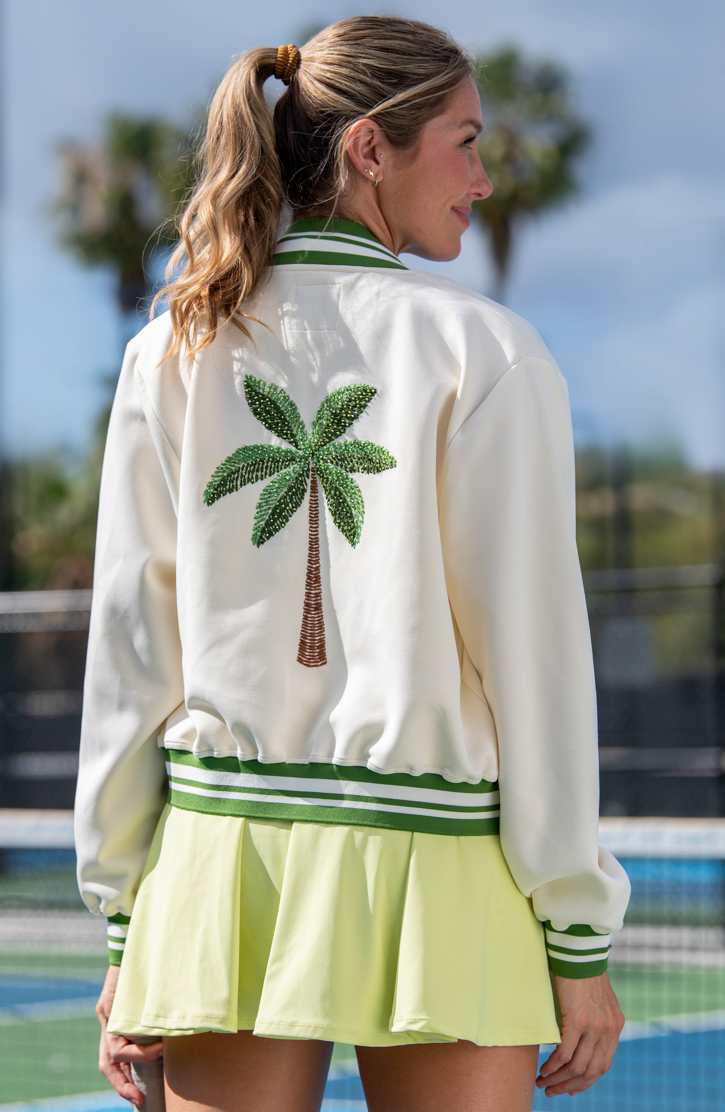 Woman wearing a white jacket with a palm tree design and a light green skirt by a pool.