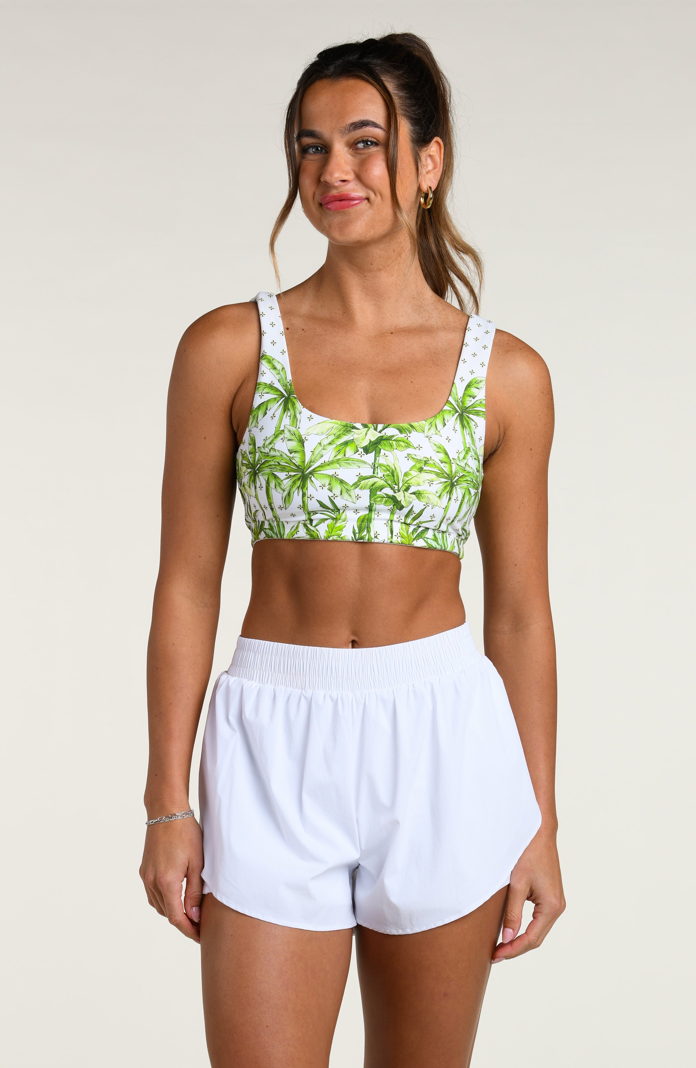 Woman wearing a white sports bra with green palm trees and white shorts against a white background. 