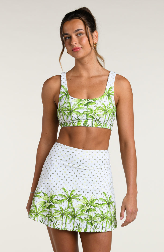 Woman wearing a white sports bra with green palm trees and a matching skort against a white background. 