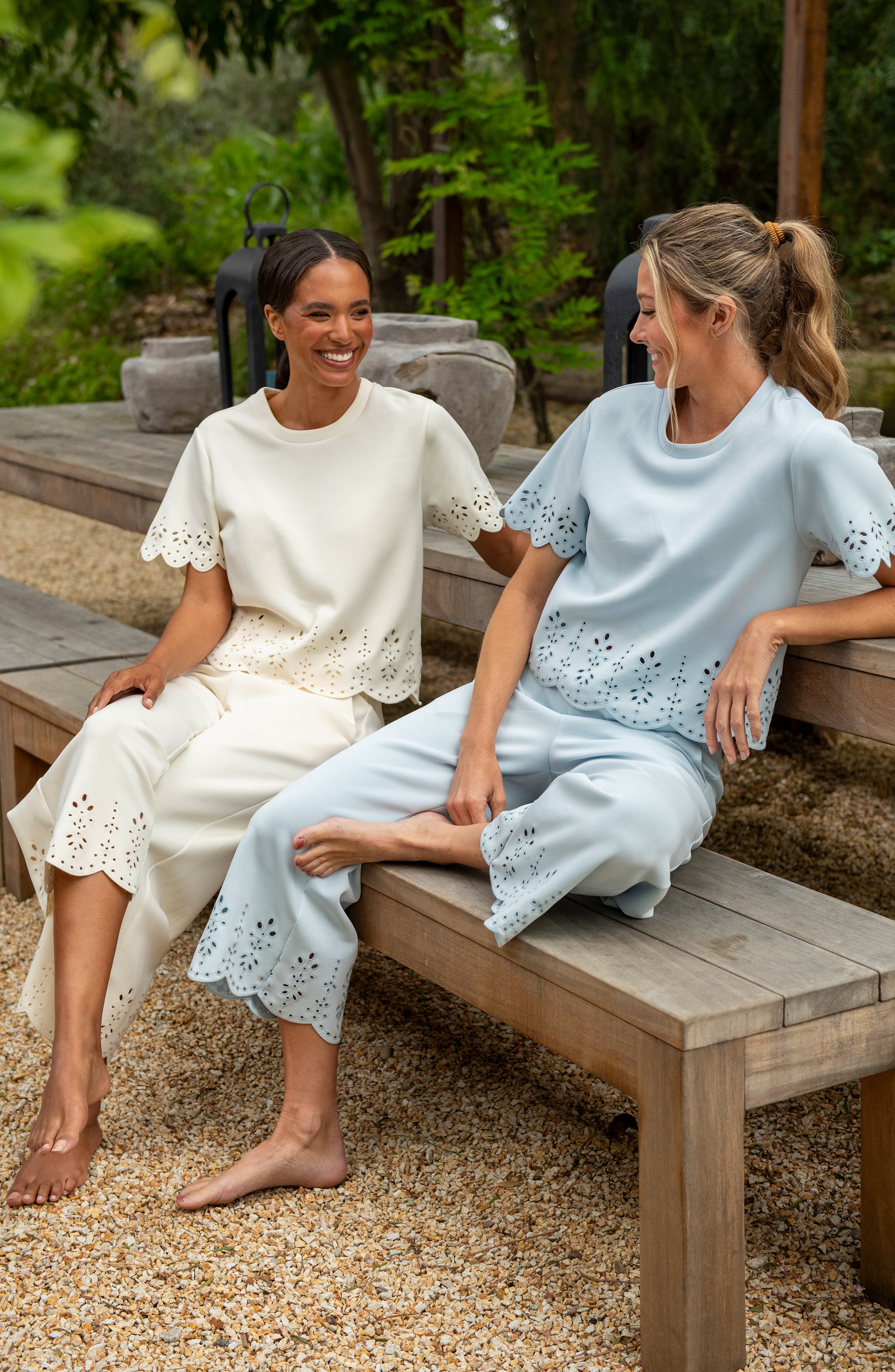 Two women sitting on a wooden bench outdoors, wearing light-colored outfits.
