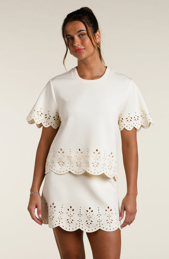 Woman wearing a beige short sleeve top with scalloped edges and a matching skirt on a plain background