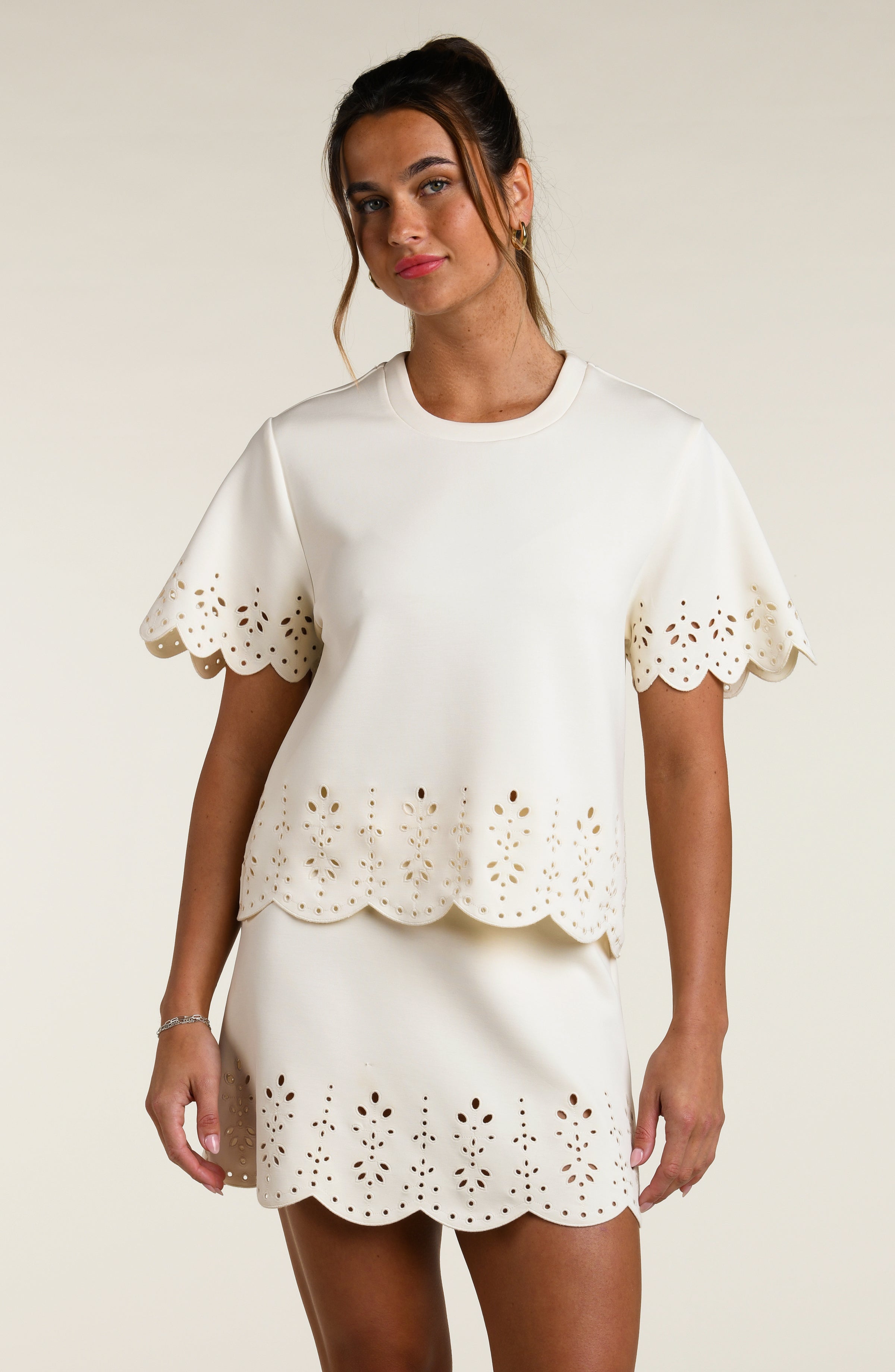 Woman wearing a beige short sleeve top with scalloped edges and a matching skirt on a plain background