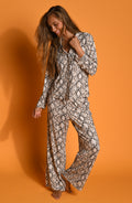 A woman modeling a matching long-sleeve button-up pajama shirt and wide-leg pants set with an all-over black and beige snakeskin pattern, shot against an orange studio background.