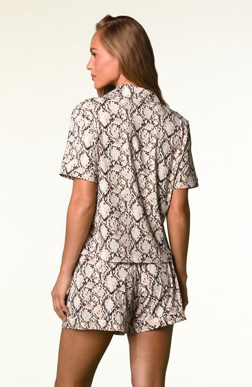 A woman modeling a matching short-sleeve button-up shirt and shorts pajama set with a black and beige snakeskin pattern, shot against a plain white studio background.