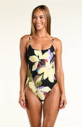 A woman modeling a one-piece swimsuit with a black background and a large floral print in pale yellow, purple, and green, photographed against a plain light background.