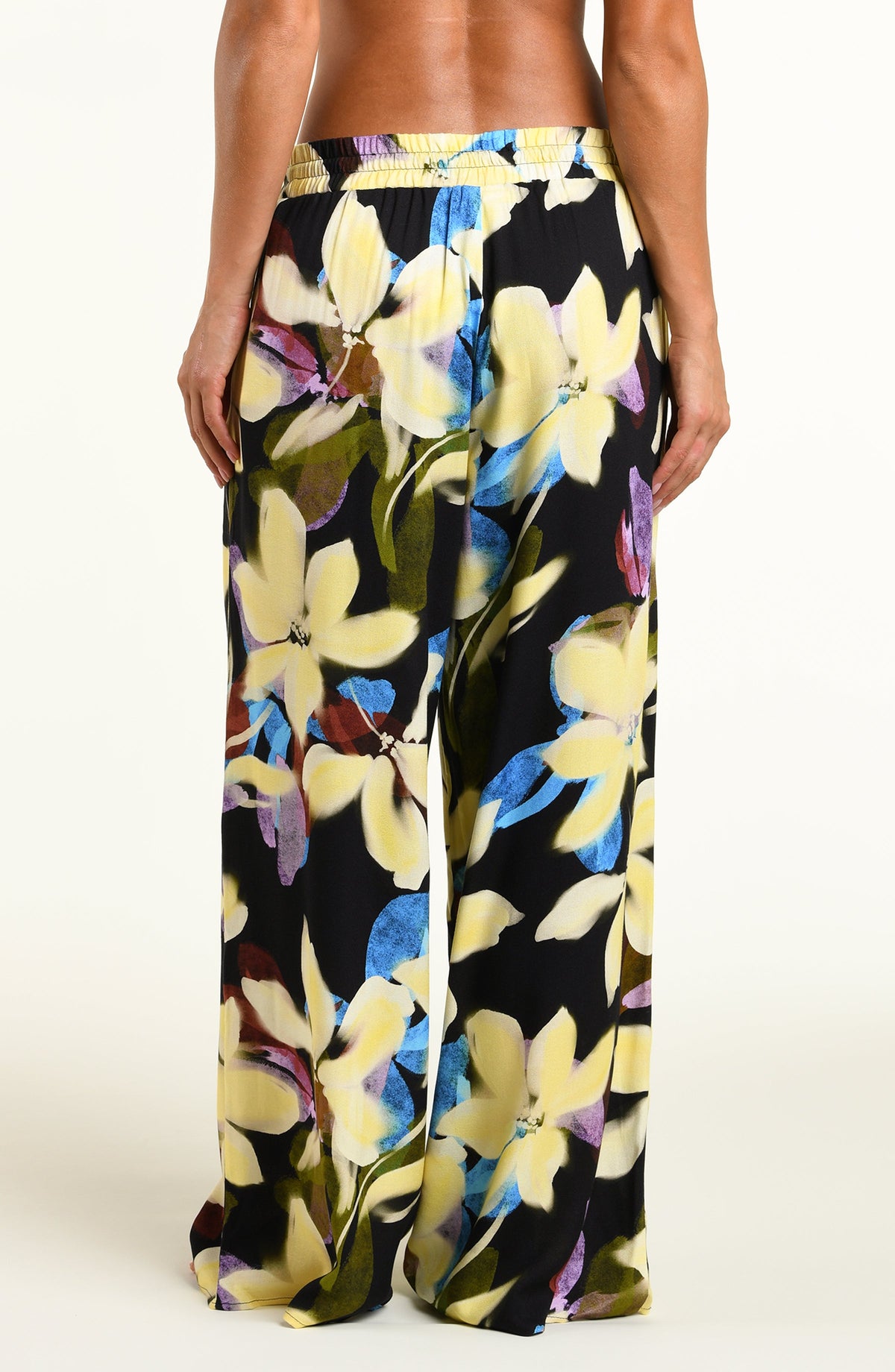A model wearing wide-leg black pants with a vibrant yellow, blue, and purple floral print, standing against a plain white studio background.