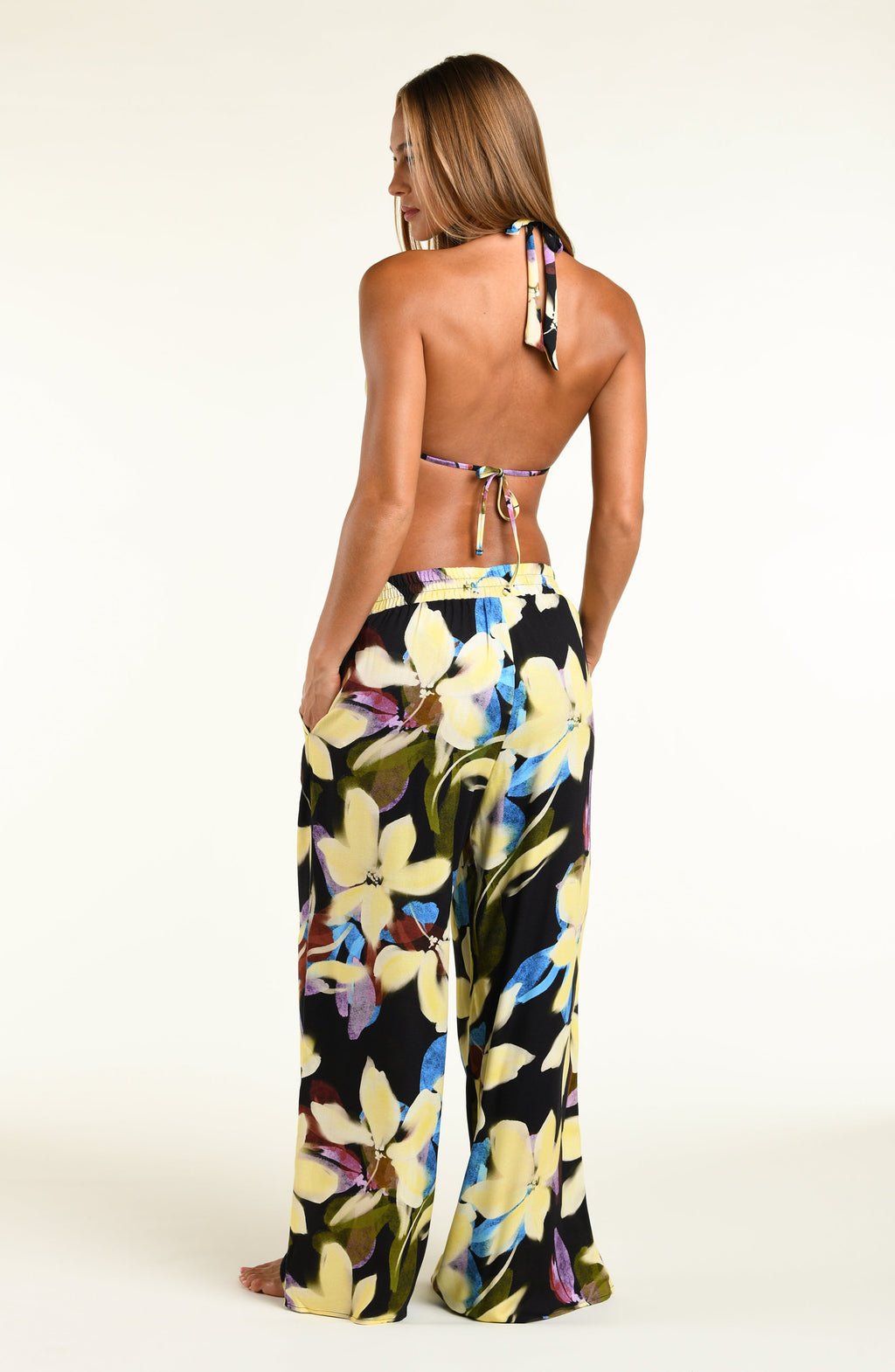 A model wearing a bikini top and matching beach pants with a vibrant yellow, blue, and purple floral print, standing against a plain white studio background.