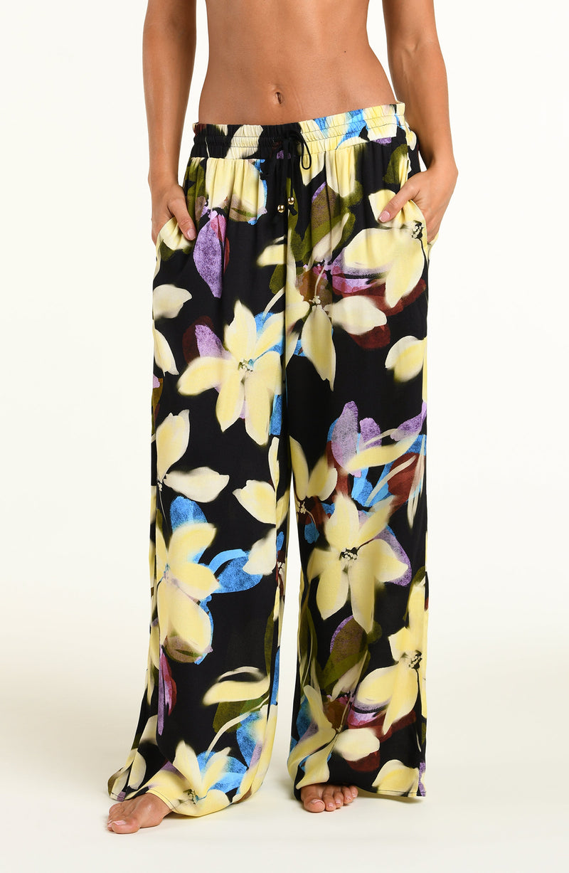 A model wearing wide-leg black pants with a vibrant yellow, blue, and purple floral print, standing against a plain white studio background.