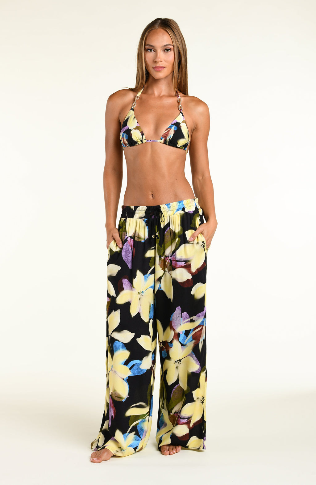 A model wearing a bikini top and matching beach pants with a vibrant yellow, blue, and purple floral print, standing against a plain white studio background.