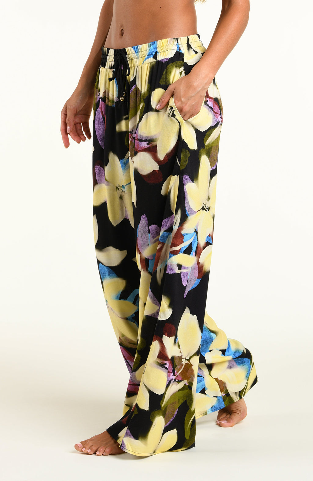 A model wearing wide-leg black pants with a vibrant yellow, blue, and purple floral print, standing against a plain white studio background.