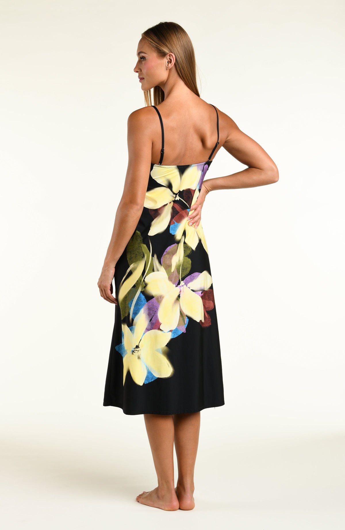 A woman modeling a black midi dress with removable straps and a large, vibrant tropical floral print in yellow, purple, and green, set against a plain off-white studio background.