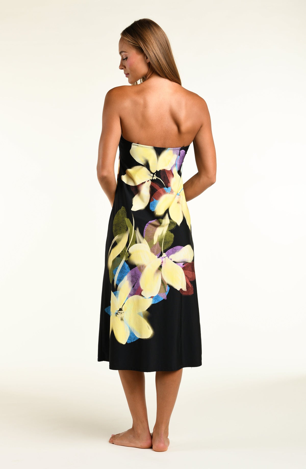 A woman modeling a black midi dress with removable straps and a large, vibrant tropical floral print in yellow, purple, and green, set against a plain off-white studio background.