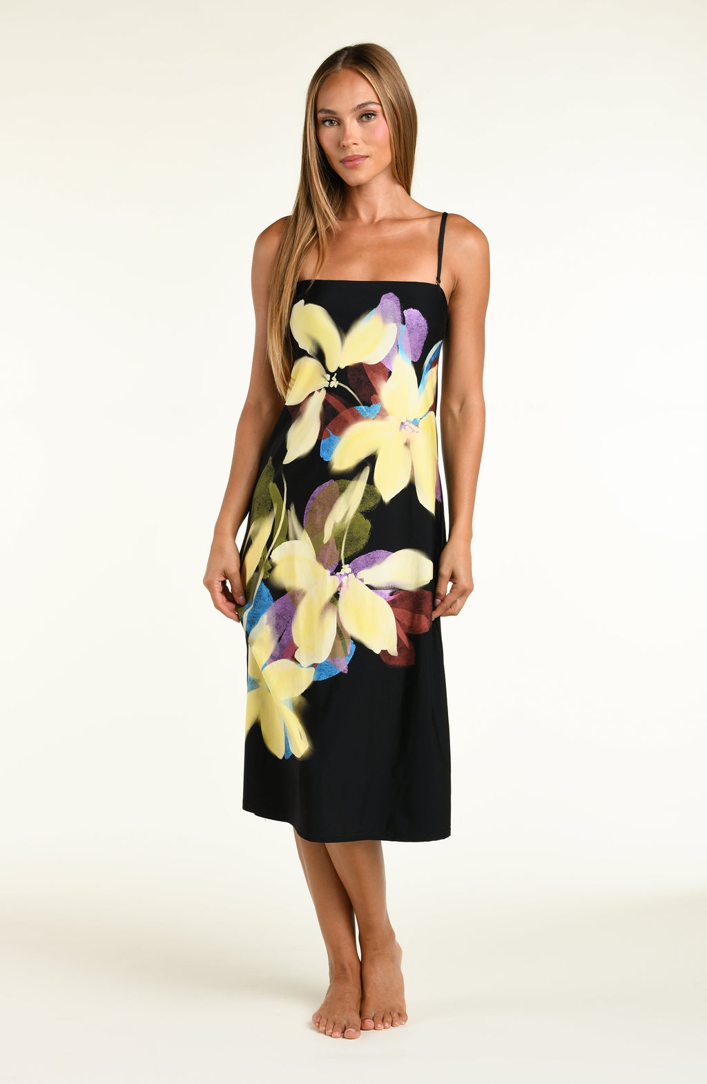 A woman modeling a black midi dress with removable straps and a large, vibrant tropical floral print in yellow, purple, and green, set against a plain off-white studio background.