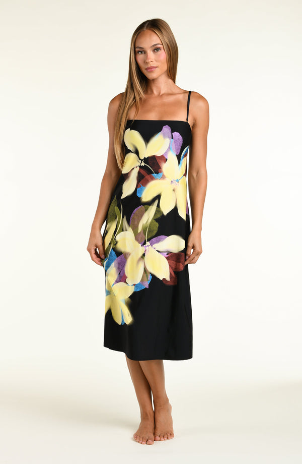 A woman modeling a black midi dress with removable straps and a large, vibrant tropical floral print in yellow, purple, and green, set against a plain off-white studio background.