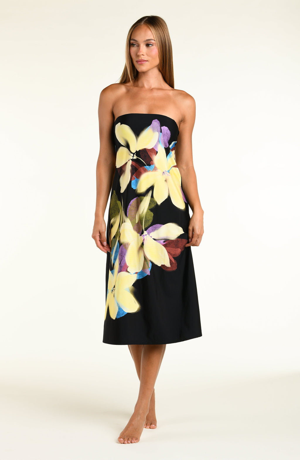 A woman modeling a black midi dress with removable straps and a large, vibrant tropical floral print in yellow, purple, and green, set against a plain off-white studio background.