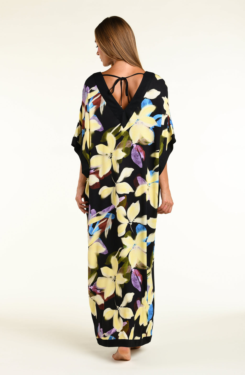 A woman modeling a black maxi caftan with a large yellow, green, and blue floral print, set against a plain off-white studio background.