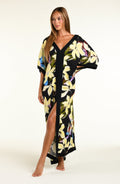 A woman modeling a black maxi caftan with a large yellow, green, and blue floral print, set against a plain off-white studio background.