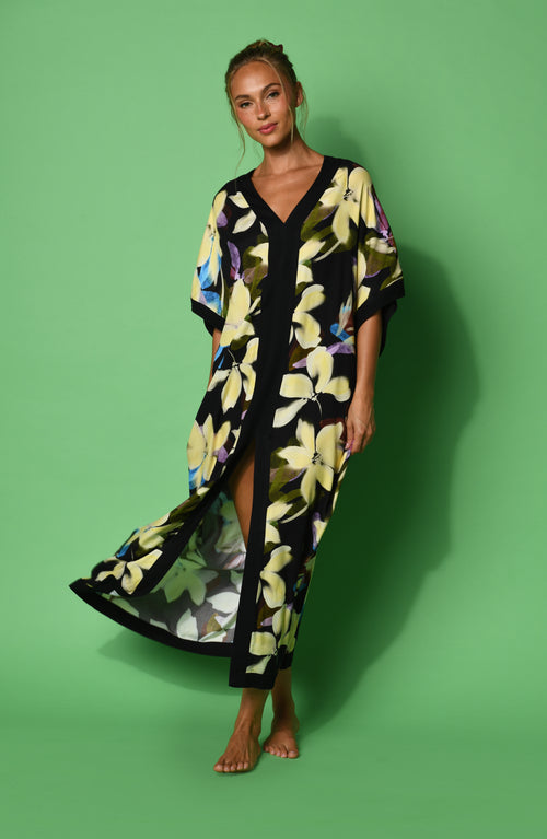 A woman modeling a black maxi caftan with a large yellow, green, and blue floral print, set against a green background.