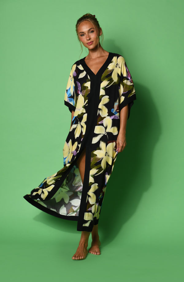 A woman modeling a black maxi caftan with a large yellow, green, and blue floral print, set against a green background.