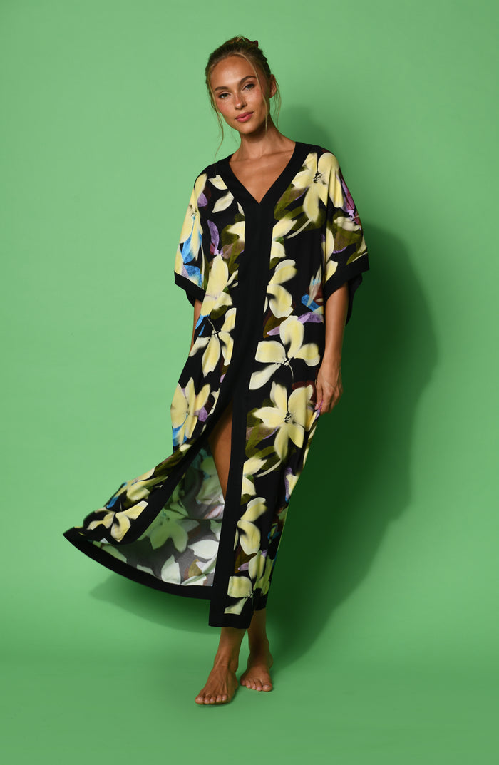 A woman modeling a black maxi caftan with a large yellow, green, and blue floral print, set against a green background.