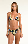 A woman modeling a two-piece bikini with a black background and a vibrant yellow, blue, and white tropical floral print, set against a plain light beige studio background.