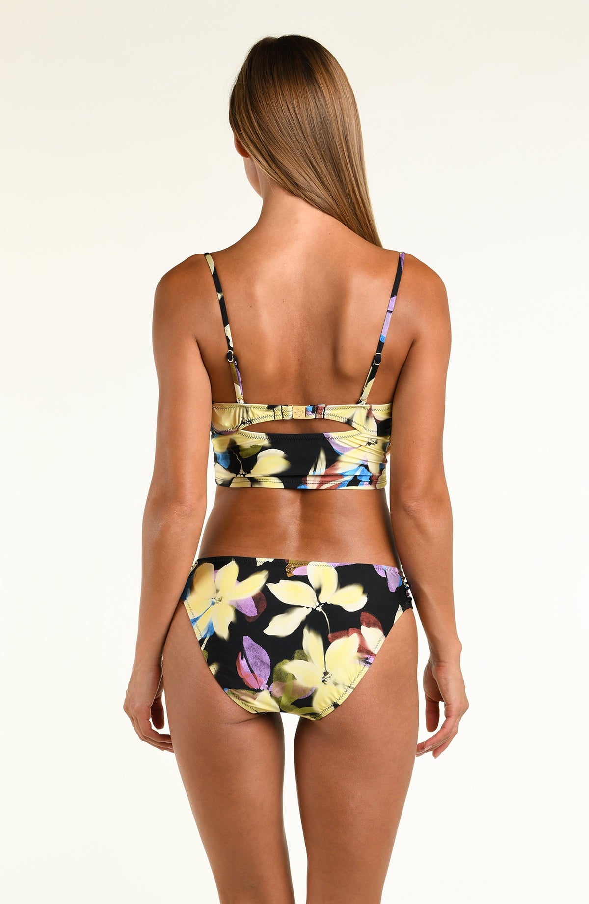 A woman modeling a two-piece bikini set with a vibrant multicolor floral print on a black background, photographed against a solid off-white studio background.