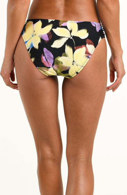 A model wearing a pair of bikini bottoms with a black background and a yellow, purple, and white floral print, shown against a plain white studio background.