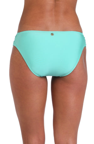 Model is wearing a solid pale, greenish-blue (seaglass) colored side shirred hipster swimsuit bottom.