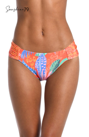 Model is wearing an orange multicolored tropical printed reversible side shirred hipster bikini bottom from our Sunshine 79 brand.
