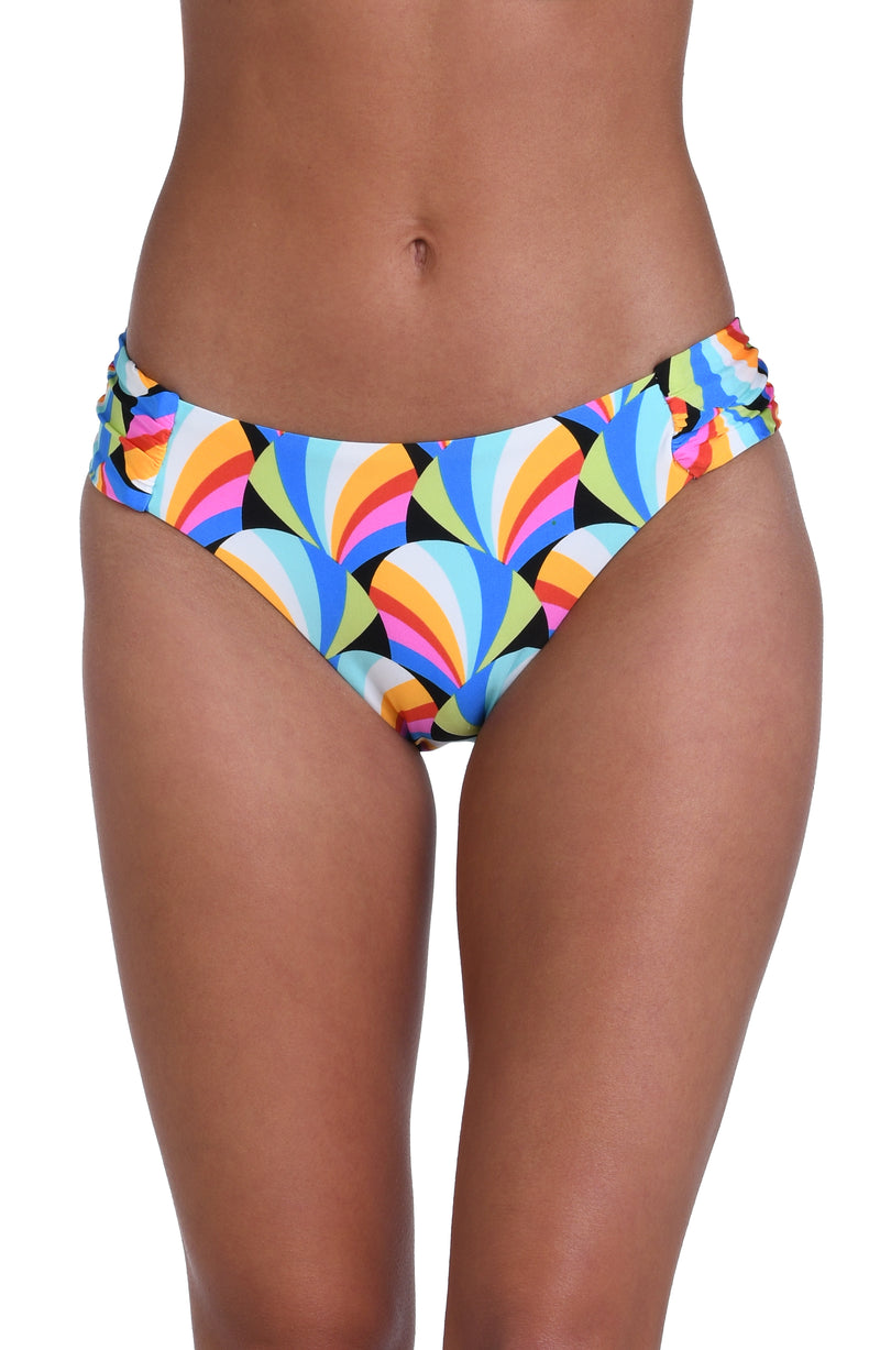 Model is wearing a pink, blue, yellow, and orange multicolored side shirred hipster bikini bottom on a geometric patterned black background.