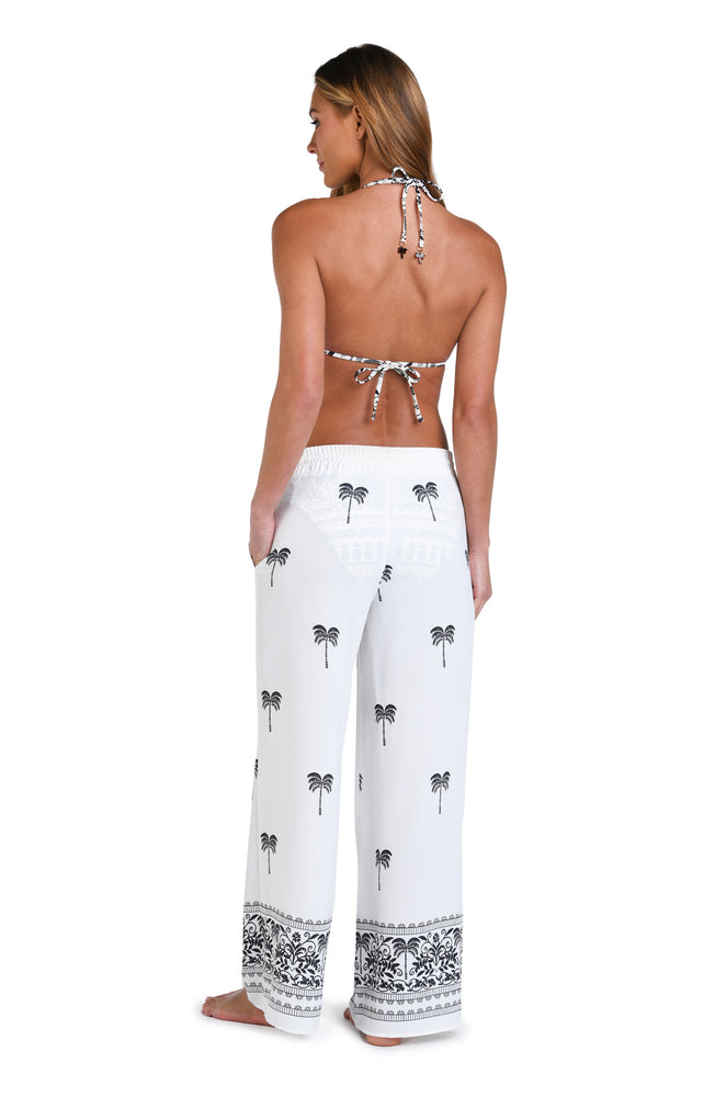 Model is wearing a black and white monochromatic patterned beach pants that features an allover tropical motif with hints of palm trees and pineapples set against a black background.