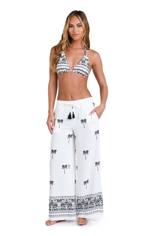 Model is wearing a black and white monochromatic patterned beach pants that features an allover tropical motif with hints of palm trees and pineapples set against a black background.