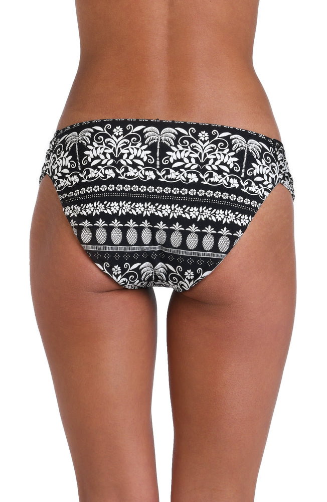 Model is wearing the reversible black and white monochromatic patterned side shirred hipster bottom that features an allover tropical motif with hints of palm trees and pineapples set against a black background.