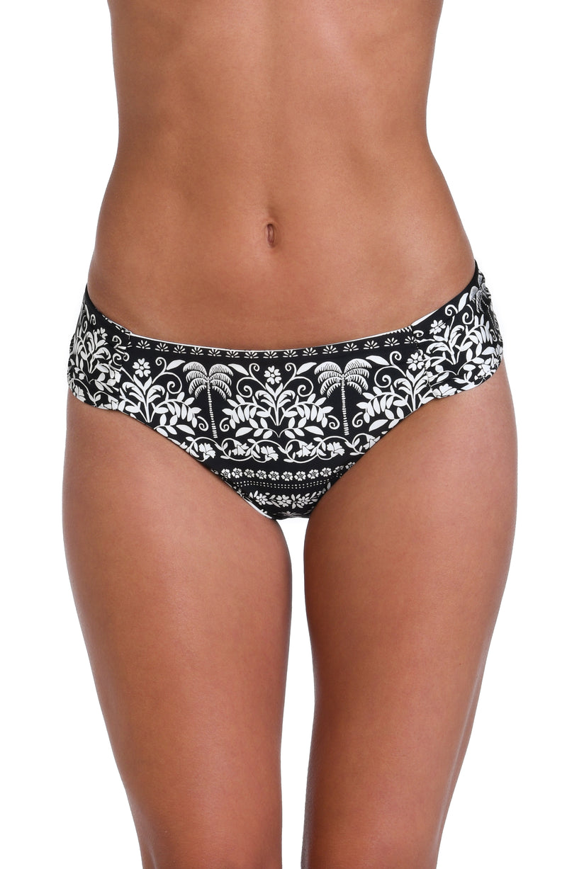 Model is wearing the reversible black and white monochromatic patterned side shirred hipster bottom that features an allover tropical motif with hints of palm trees and pineapples set against a black background.