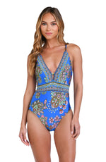 Model is wearing a pink, orange, teal, and green multicolored floral patterned plunge one piece swimsuit set against an indigo blue background.