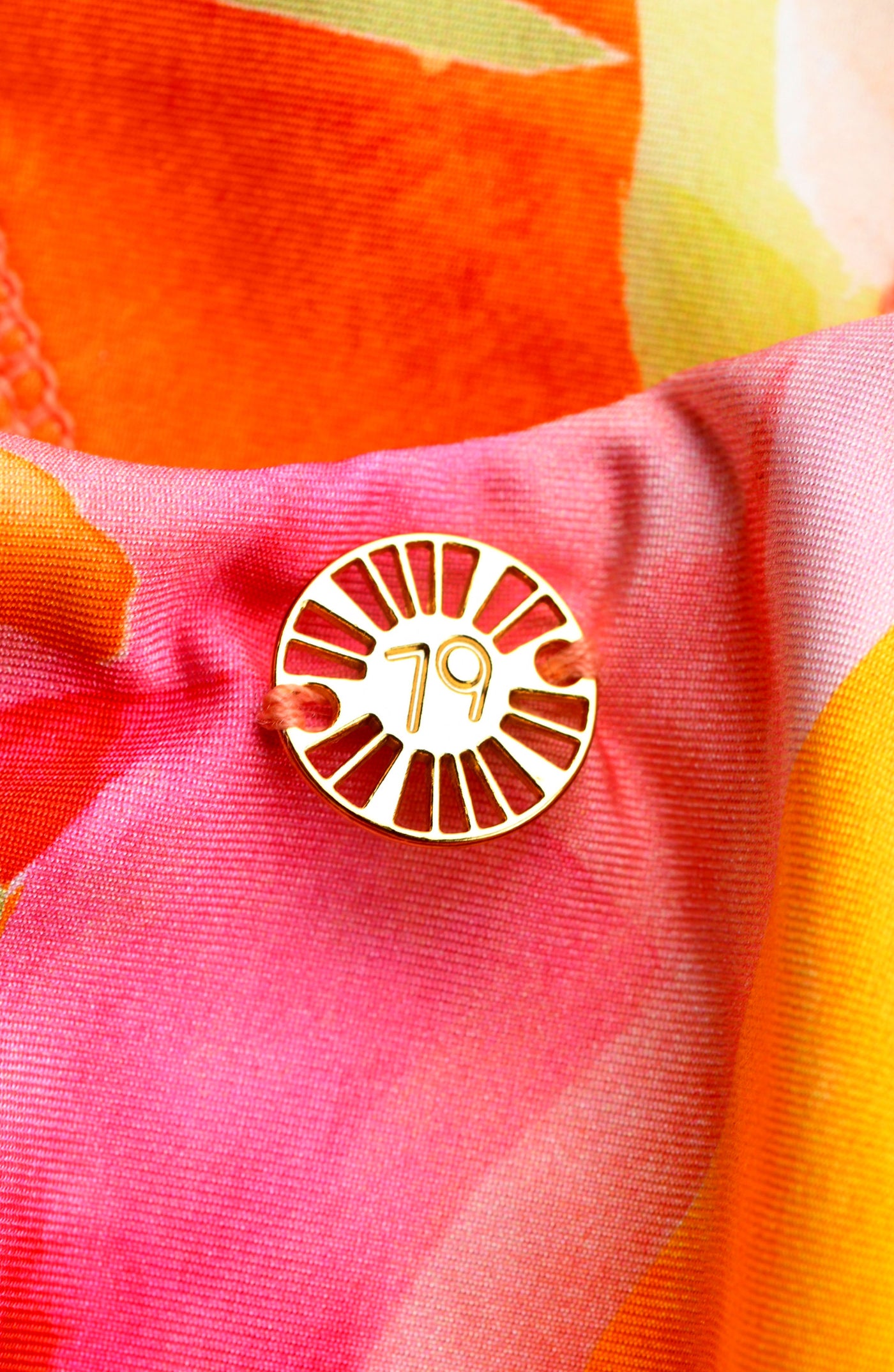 
                  
                    Close-up of a gold front charm on a multicolored tropical printed one piece swimsuit. 
                  
                