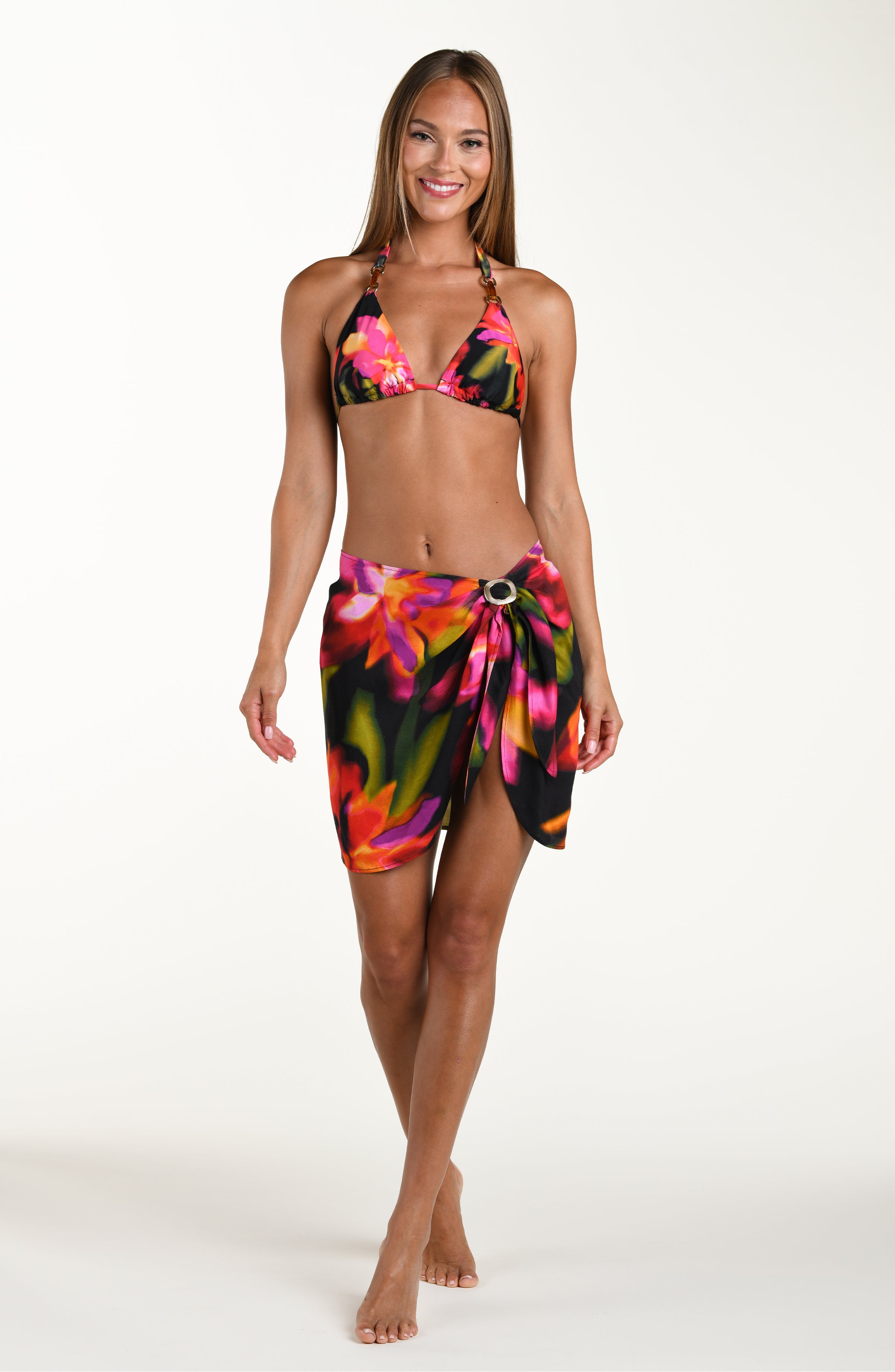 Full body front image of model wearing a short pareo cover up with a gold slider, featuring a bright floral print. This is paired with a matching bikini top and bottom.