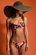 Lifestyle image of model wearing a bandeau bikini top with a removable halter strap, twist front design, and gold ring accent at the center, featuring a multicolored floral print. This is paired with matching bikini bottoms.