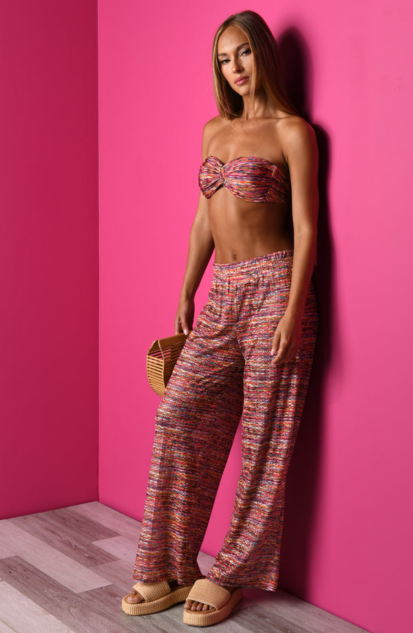 Lifestyle image of a bandeau bikini top in a multicolored ripple pattern, featuring a ruched center with a ring detail and removable halter strap.